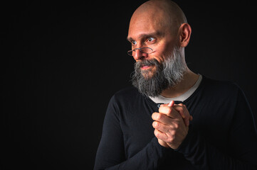 Funny bearded man with mischievous expression and clasped hands. Bald adult male posing on black background. Concept of secret plan or cunning idea