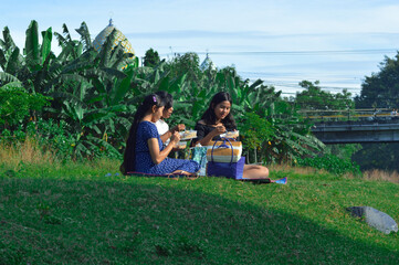 Three teenage girls savor their food in the calm outdoors, sitting comfortably on a picnic mat...