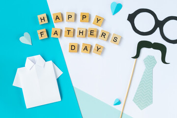 Flat lay with Happy Fathers Day message paper shirt blue tie glasses mustache wooden letters blue hearts on white and blue background celebration photo for dad gift theme