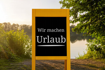 Aufsteller mit der Aufschrift WIR MACHEN URLAUB vor dem Motiv eines Sommerabends am See