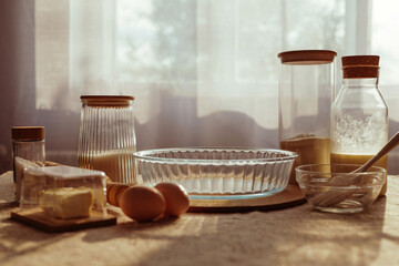 A tabletop still life features ingredients for baking, including flour in glass jars, eggs, butter, and a glass baking dish, illuminated by window light.