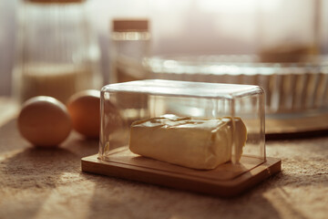 A close-up focuses on a stick of butter in a glass dish with a wooden base, alongside two brown eggs, on a textured surface with blurred baking items in the background.