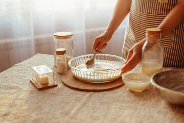 A cook in a striped apron spreads batter in a glass pie dish with a spoon, surrounded by baking...