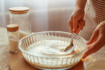 A cook in a striped apron spreads batter evenly in a round glass baking dish with a spoon, with jars of ingredients nearby.
