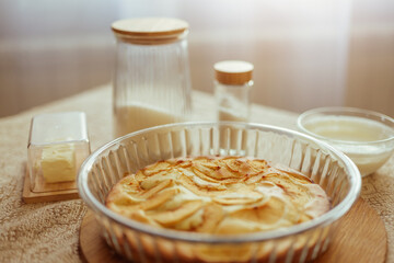 A baked apple pie sits in a glass dish on a wooden board, surrounded by baking ingredients like...