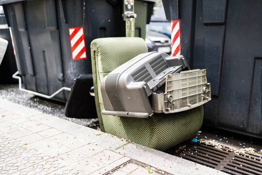 Broken television and chair abandoned near dumpsters on sidewalk