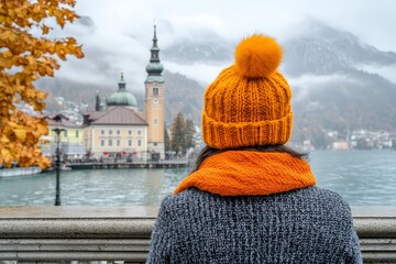 View of a woman wearing an orange hat and scarf enjoying the autumn scenery in a lakeside town with mountains in the background