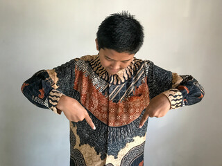 A teenage boy wearing a batik shirt is pointing both of his index fingers downward, isolated on white