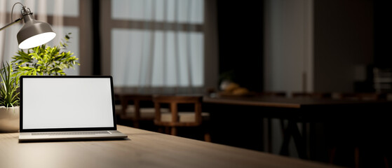 White screen laptop on wooden table under lamp light in dim lit living room or dining room.