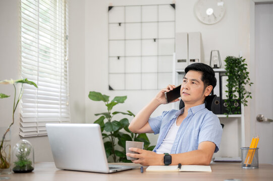 Asian man holding coffee mug and talking on phone at wooden table in his office or working room.