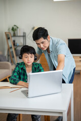 Fototapeta premium Asian father teaching kid elementary school boy wearing headphones while looking at laptop together.