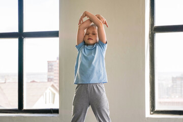 A child performs a yoga pose on a mat indoors, enjoying natural light streaming through large...