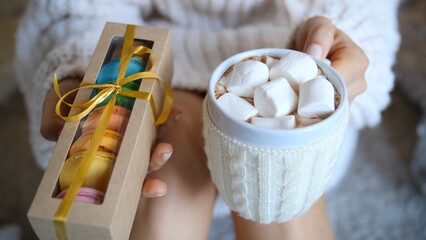 Woman wearing a white sweater holding a mug of hot chocolate with marshmallows and a gift box of...