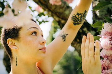 Tattooed woman reaches toward pink cherry blossoms in sunlight