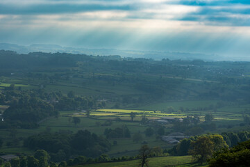 Sunbeams breaking through clouds over the patchwork fields of Wensleydale, Yorkshire Dales, England – rural countryside landscape with dramatic morning light and misty layers