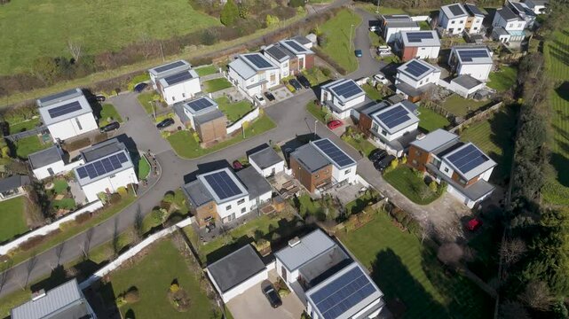 High aerial of broad estate rows of detached solar houses in varied layouts, white-grey-brown facades and private gardens expressing estate-wide sustainability