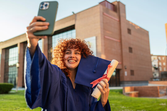 A student in a graduation gown takes a selfie on campus, celebrating her achievement.