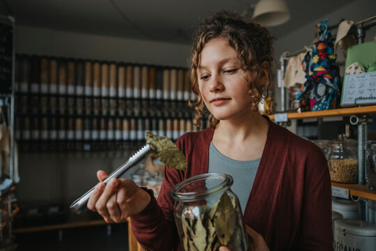 Young female store owner looking at bay leaf while holding with tong in retail shop