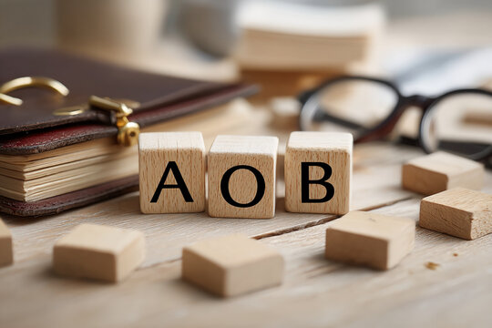 Wooden cubes spelling AOB on light wood table, near book and glasses, suggesting a business or planning concept