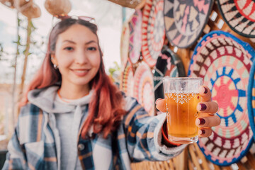 Young woman holding a glass of traditional moroccan tea, with colorful handmade souvenirs in the...