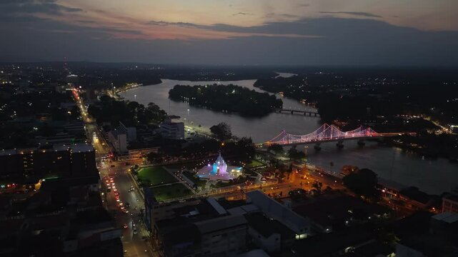 Night aerial of Surat Thani City Pillar Shrine glowing, with city lights, Tapi River, and traffic illuminating the urban landscape of Thailand.
