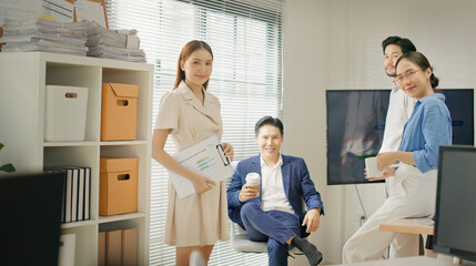 Diverse business professionals posing casually and confidently in bright modern office. Group of Asian coworkers taking a break, holding coffee mugs and smiling, promoting teamwork and positivity.