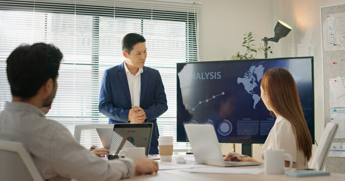 Asian executive presents business data and growth analytics on screen to diverse corporate team during strategy meeting. Multinational group focuses on digital performance, sales, and planning.