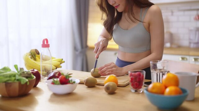 Healthy Asian woman in activewear prepares a fresh fruit drink, slicing fruit and blending a protein shake in her modern pantry kitchen at home, promoting wellness and fit lifestyle.