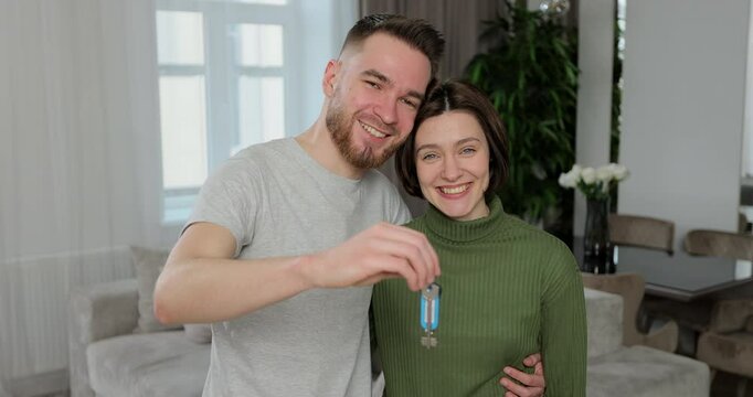 Young couple first time buying house receiving hugging looking at camera holding bunch of keys. Portrait of laughing spouses real estate owners in new apartment.