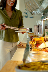 Smiling woman serving cheese with wooden tongs at a breakfast buffet in a hotel restaurant, enjoying a delightful morning meal