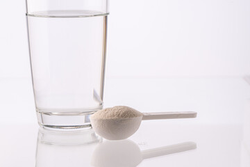 Collagen powder in a measuring spoon and a glass of water on a white background with reflection. Medicinal powder prepared for ingestion or dissolution in water.