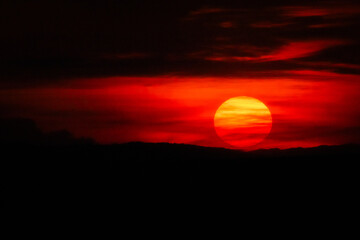 A centered sun radiates through a fiery red sunset, framed by dark hills. Perfect for bold nature...