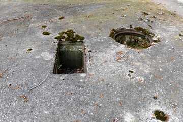 Weathered concrete ground with two open, moss-covered holes, surrounded by debris and signs of...