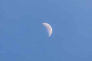 A sharp crescent moon floats in a pure blue sky, captured in high clarity during daylight hours. A...