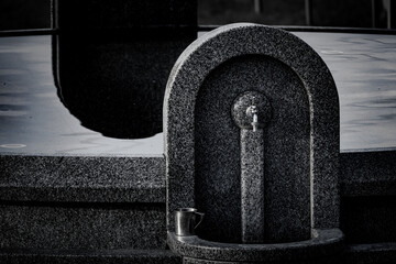 Close-up of a granite water fountain with a chrome tap and metal mug. Stone craftsmanship and still water in religious or public setting
