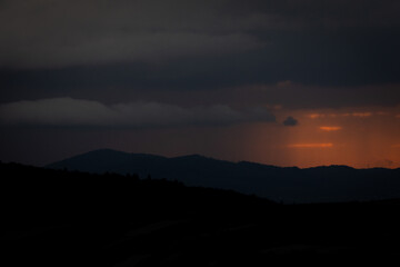 Dark mountain silhouettes under a dramatic sky with streaks of orange light at sunset. A perfect blend of contrast and calmness in a moody natural landscape