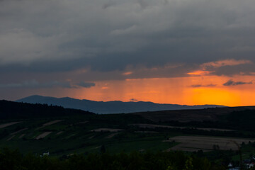 Vibrant sunset light pierces through layers of clouds over farmland and hills, casting long shadows and painting the sky with warm tones. A captivating rural moment.