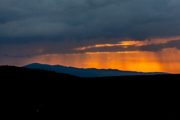 Dramatic orange twilight glows behind a mountainous horizon as storm clouds roll in, casting vertical rain curtains against the golden dusk sky — a powerful landscape full of contrast and emotion