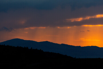 A fiery sunset breaks through dark clouds, painting the horizon in vibrant hues of orange and red while light rain gently falls over distant mountain ranges — a scene of serene wilderness