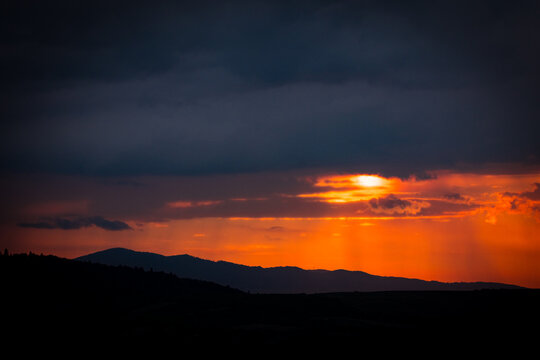 The last light of day filters through heavy clouds and showers, casting radiant streaks across a brooding sky above a layered mountainous backdrop — nature’s quiet yet powerful spectacle