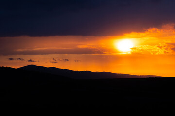 A radiant sunset over layered hills with a glowing orb descending behind cloud lines