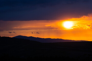 Intense sun framed by clouds as it sets beyond a forested mountain range.