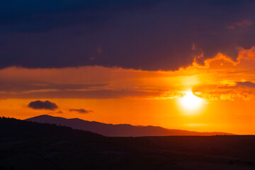 Evening sun blazes above forested ridges, illuminating thick clouds with fiery highlights and a deep amber sky