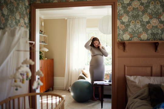Young pregnant woman talking on smart phone while standing near fitness ball at home
