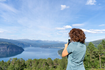 Young woman in her 30's seen from behind, capturing a stunning Norwegian of innvikfjorden fjord landscape with her mobile phone on a sunny summer day, enjoying the breathtaking scenic view