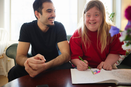 Happy woman with down syndrome coloring on book while sitting near male nurse in nursing home