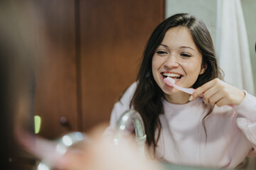 A cheerful young woman in a pastel-colored top brushing her teeth while standing before a bathroom mirror, promoting daily dental health practices and hygienic routines in a congenial setting.