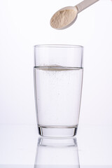 Collagen powder is poured with a measuring spoon into a glass of water on a white background with a reflection. The medicine is diluted in water. Vertical photo
