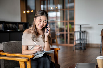 Asian woman smiling during phone call and writing in a tablet on wooden chair in cafe or coffee shop