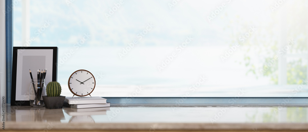 Poster a clock over books with cactus and photo frame aside on shiny marble table under wide glass window.